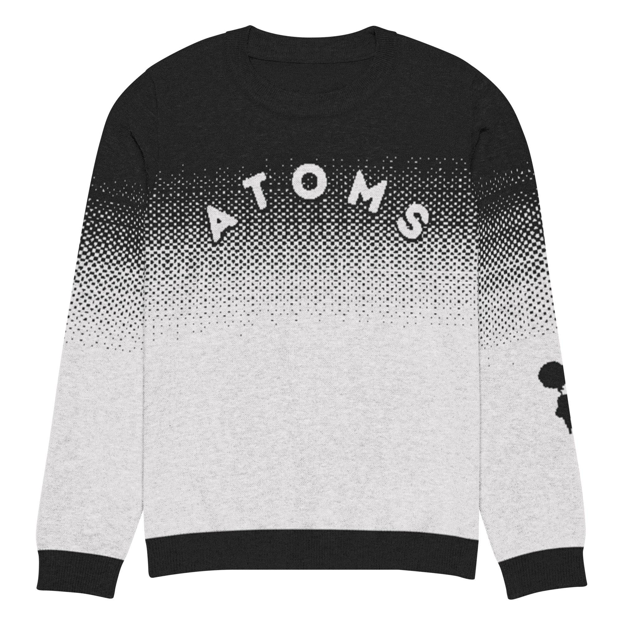 Model wearing Breakin' Atoms "Pixel Dusk Knit," a black-to-gray gradient sweater with pixelated dot fade, bold "ATOMS" typography, and parachuting silhouette graphic on the sleeve — blending cozy knitwear with streetwear flair.