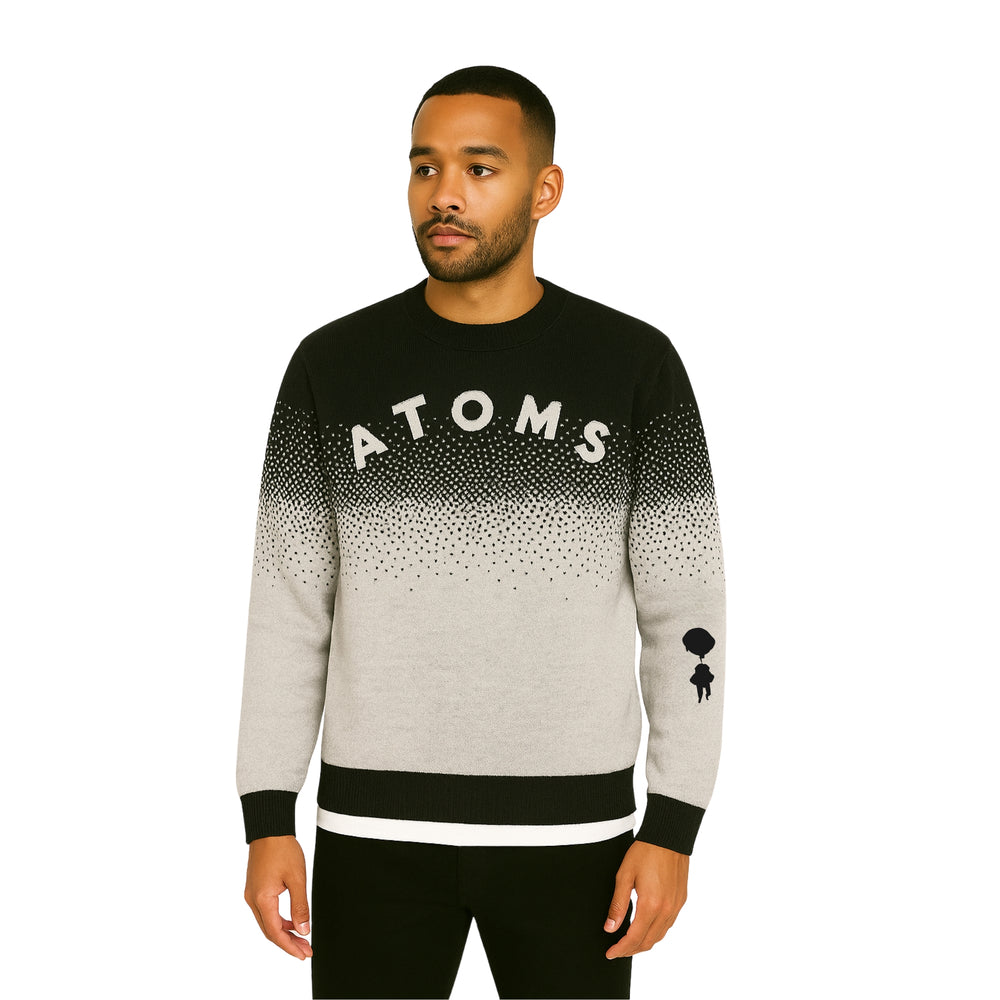 Model wearing Breakin' Atoms "Pixel Dusk Knit," a black-to-gray gradient sweater with pixelated dot fade, bold "ATOMS" typography, and parachuting silhouette graphic on the sleeve — blending cozy knitwear with streetwear flair. — Breakin Atoms premium streetwear